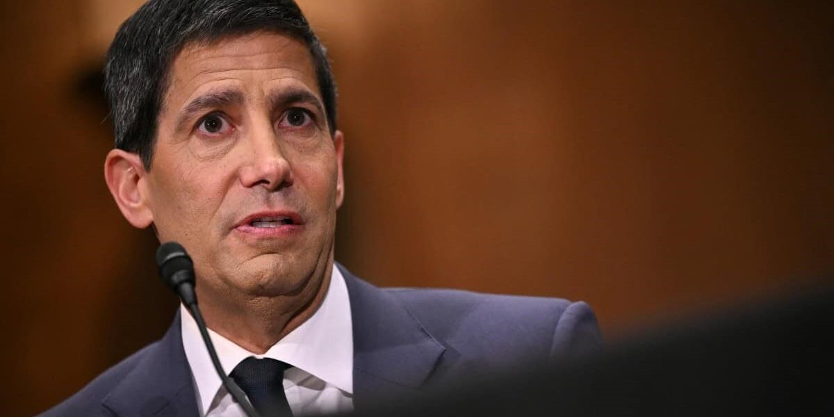 A man in a dark suit and white shirt speaks into a microphone in a formal setting, with a brown backdrop behind him.