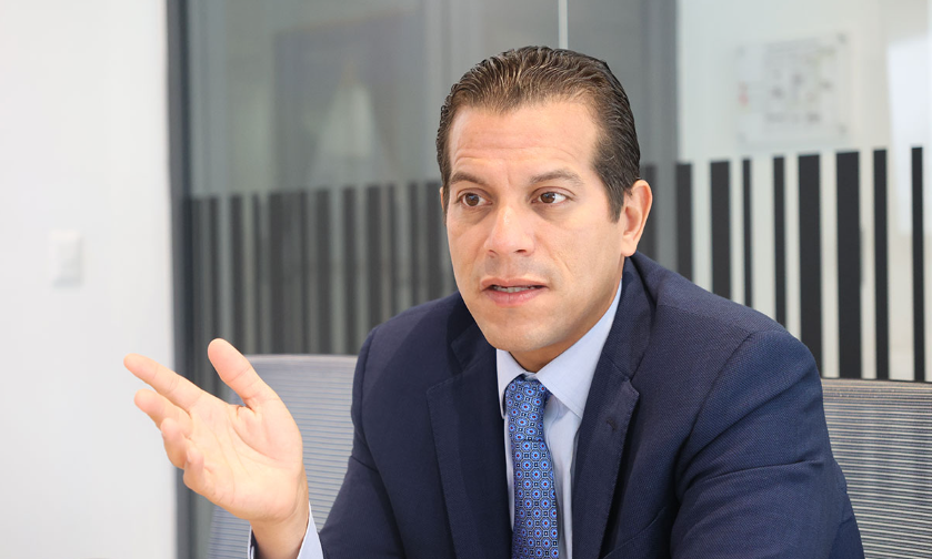 Businessman in a navy suit gestures with his left hand while speaking in a modern office setting