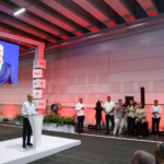 Man in a light shirt speaks at a white podium on a stage, large screen behind him, and media crew and audience observing.