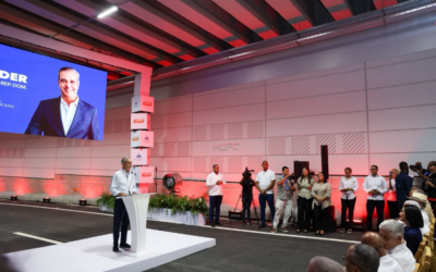 Man in a light shirt speaks at a white podium on a stage, large screen behind him, and media crew and audience observing.
