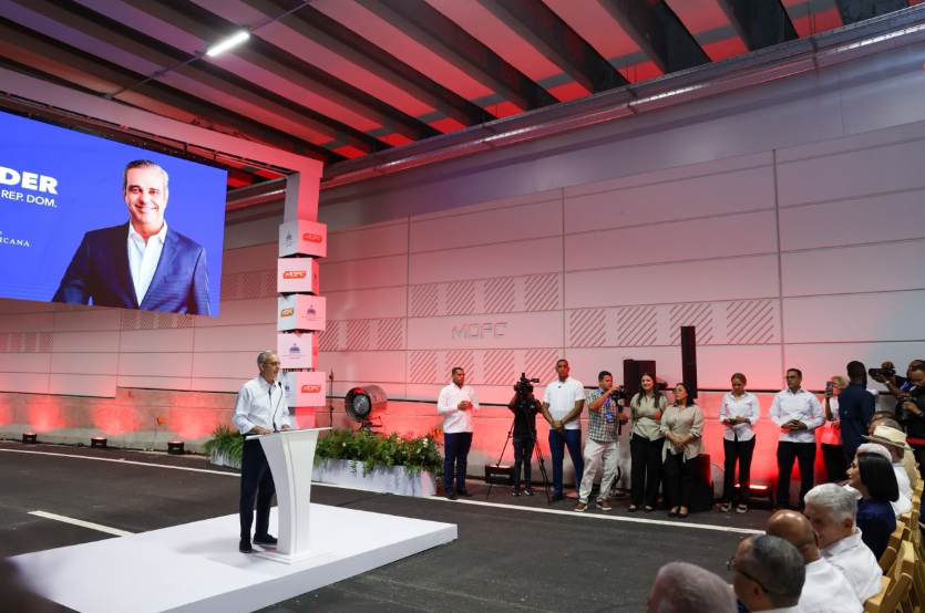 Man in a light shirt speaks at a white podium on a stage, large screen behind him, and media crew and audience observing.