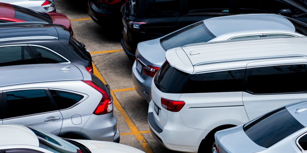 A crowded parking lot viewed from above with numerous silver and dark-colored cars parked in tight spaces, yellow lines marking spots.
