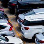 A crowded parking lot viewed from above with numerous silver and dark-colored cars parked in tight spaces, yellow lines marking spots.