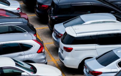 A crowded parking lot viewed from above with numerous silver and dark-colored cars parked in tight spaces, yellow lines marking spots.
