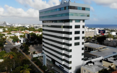 Aerial view of a tall white office building with a glass top section near the coast, surrounded by city streets and trees.
