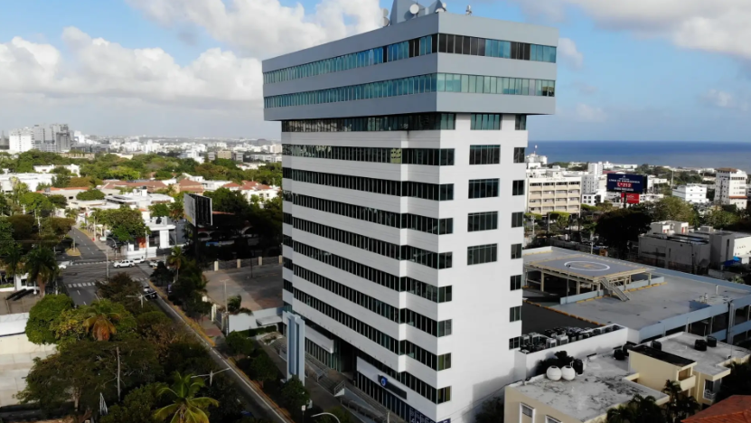 Aerial view of a tall white office building with a glass top section near the coast, surrounded by city streets and trees.