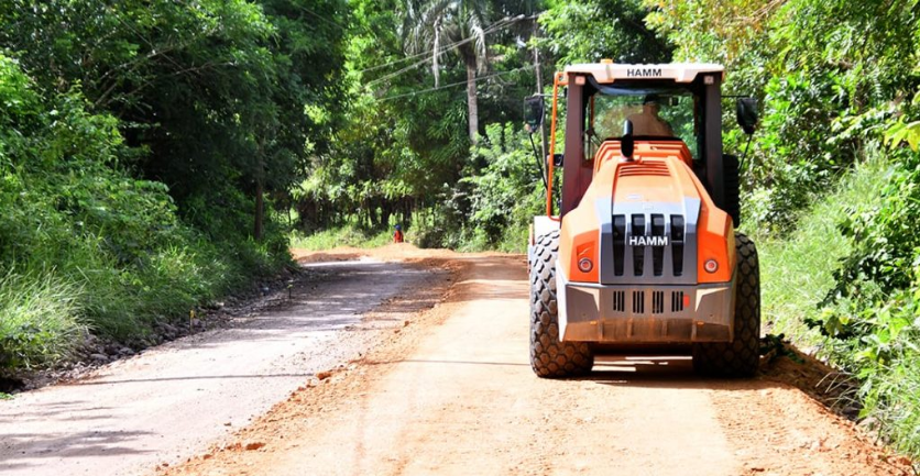 Orange road roller on a dusty dirt road flanked by dense green trees and bushes.