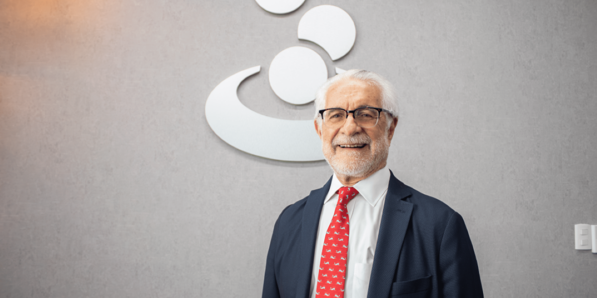 Older man in a navy suit and red tie standing indoors in front of a company logo on a gray wall, smiling for a professional portrait, business setting