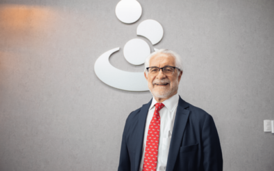 Older man in a navy suit and red tie standing indoors in front of a company logo on a gray wall, smiling for a professional portrait, business setting
